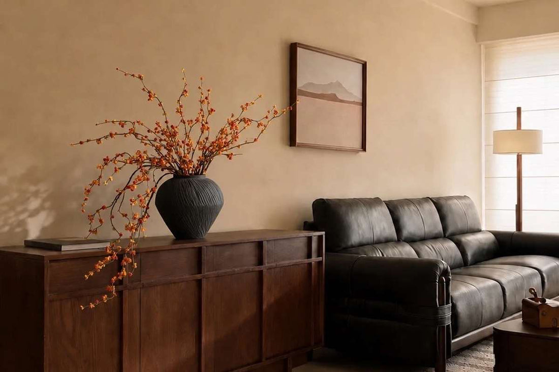 Artificial orange berry branches in black vase on wood sideboard with leather sofa for warm midcentury living room home decor