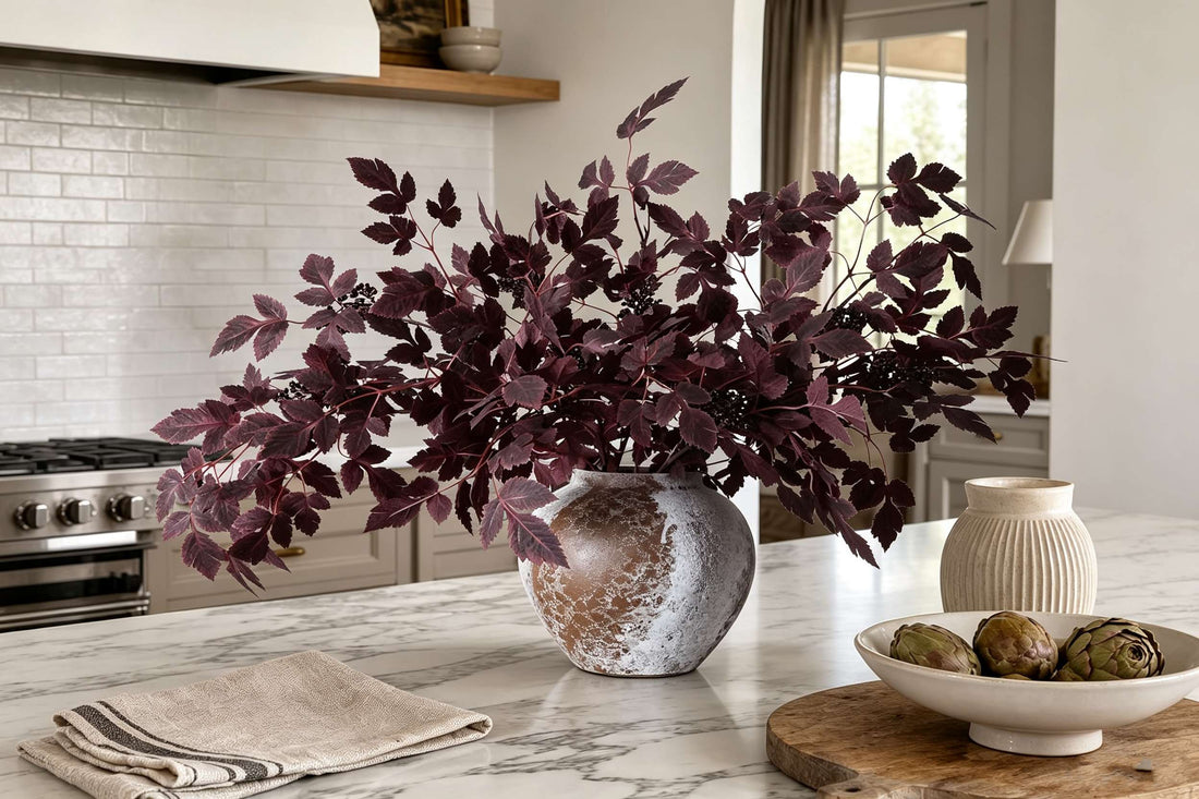 Artificial dark purple foliage stems in distressed ceramic vase styled on marble kitchen island with artichoke bowl decor