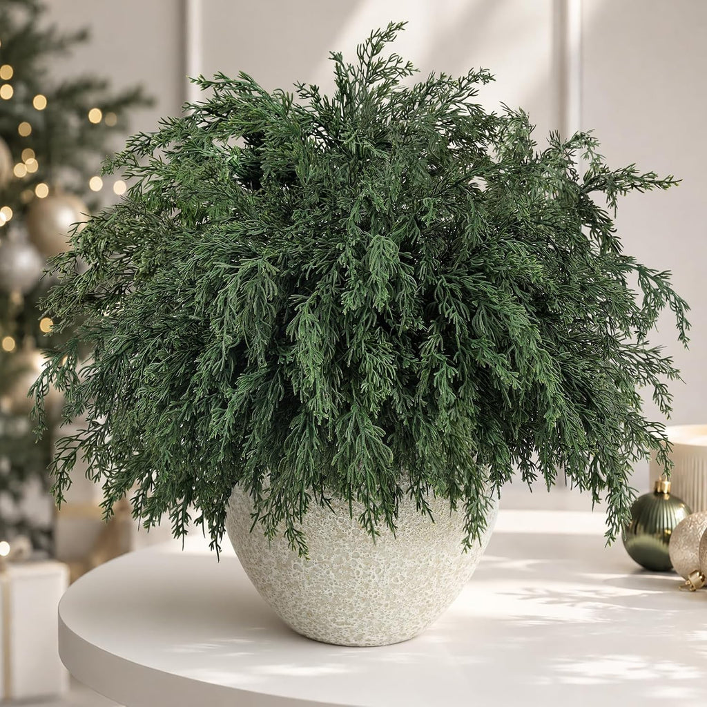 Artificial cedar pine arrangement in gray textured vase on white round table with blurred Christmas tree ornaments and gifts for festive holiday home decor
