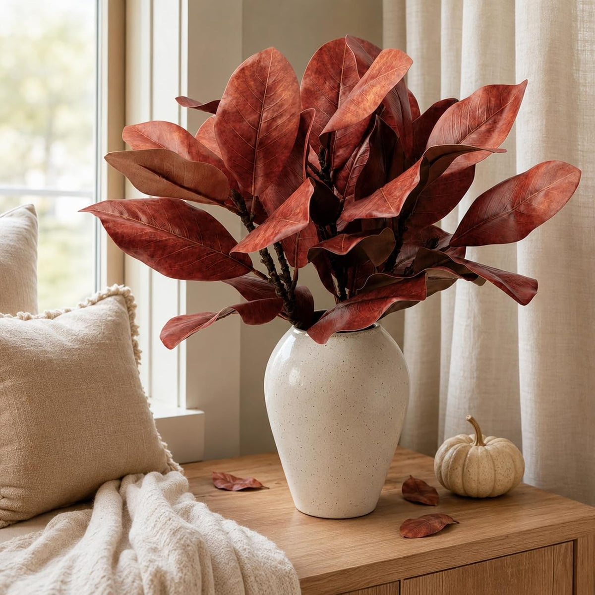 Artificial magnolia leaves bouquet in white speckled vase on wood console with pumpkin for cozy fall autumn home decor