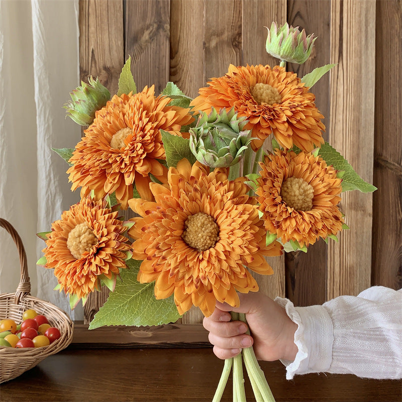 Hand holding artificial orange sunflower bouquet against rustic wood wall with wicker basket of cherry tomatoes for farmhouse country home decor style