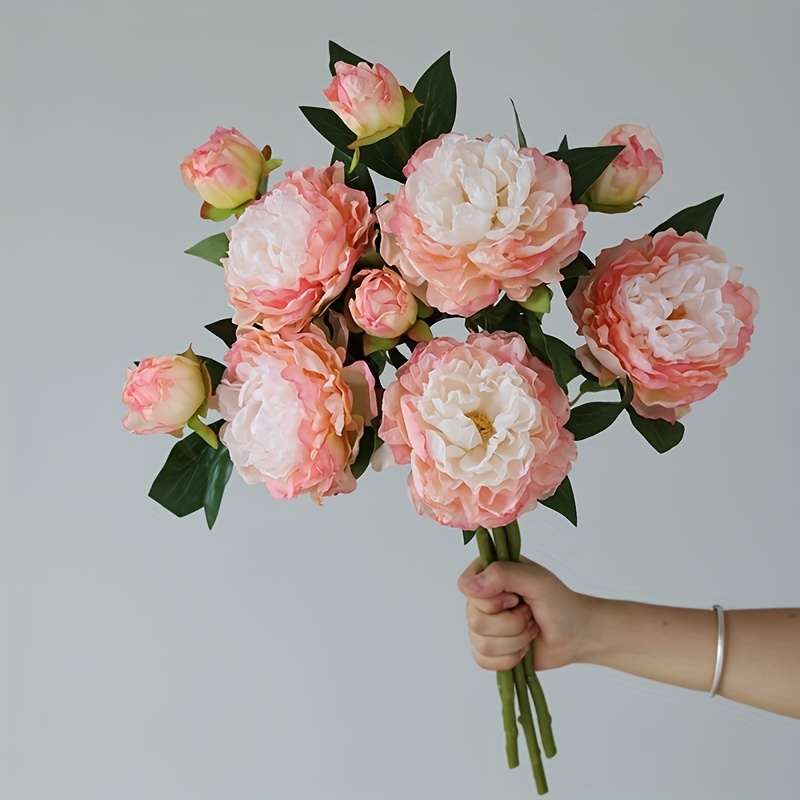 Hand holding bouquet of artificial blush pink peonies with six open blooms and six buds with green leaves against gray background showing bouquet size