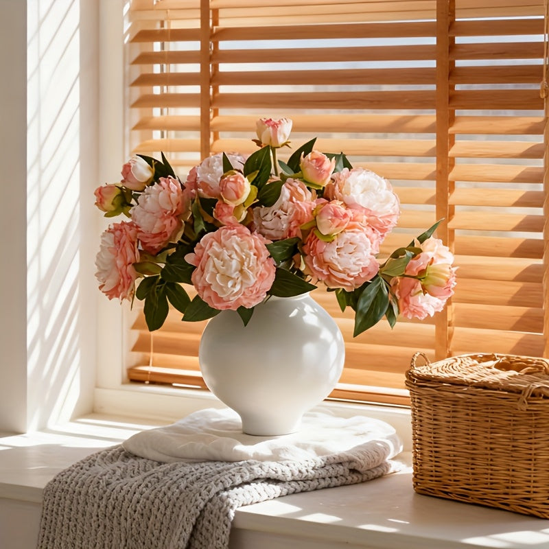Artificial blush pink peony bouquet in white vase on window sill with knit blanket and basket for cozy nordic home decor