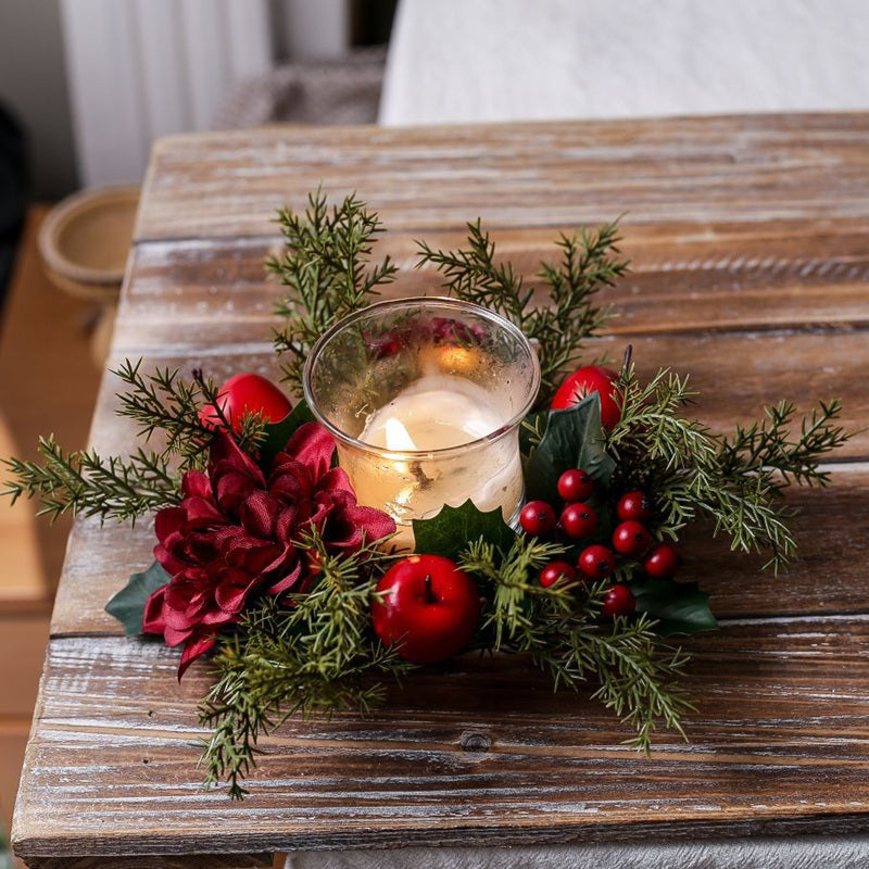 Festive Christmas candle ring featuring burgundy dahlia flower, red apples, berries and cedar leaves, lit candle on weathered wood table