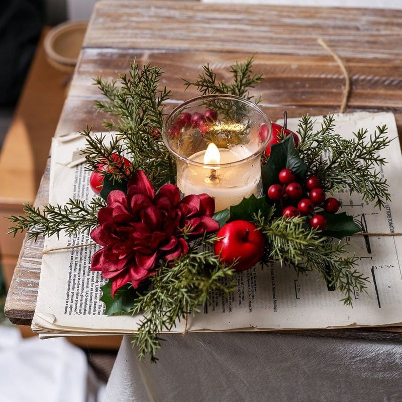 Burgundy flower Christmas candle ring with red apples, berries and cedar greenery, lit glass candle on vintage paper rustic wooden table