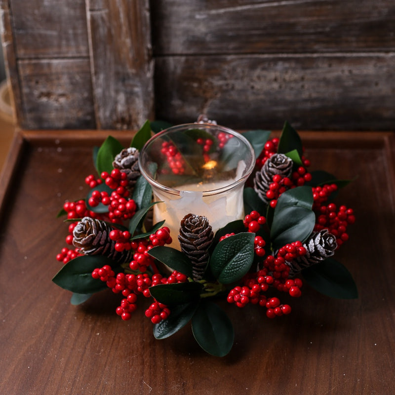 Small christmas candle ring wreath with red berries pine cones surrounding clear glass candle cup on dark wood table