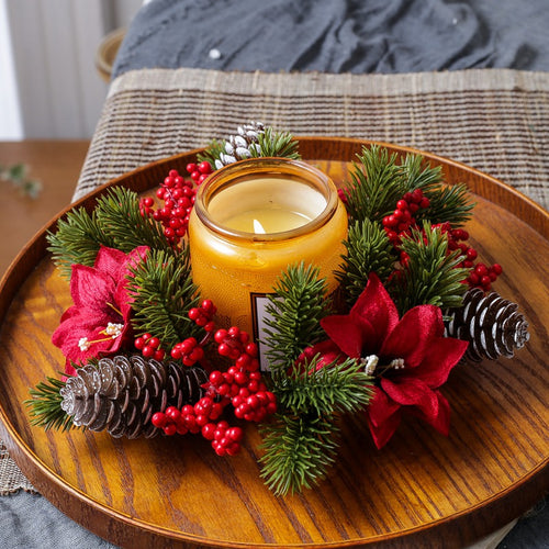Christmas candle wreath holder decorated with red poinsettia, red berries and pine greenery around amber candle jar on round wooden tray