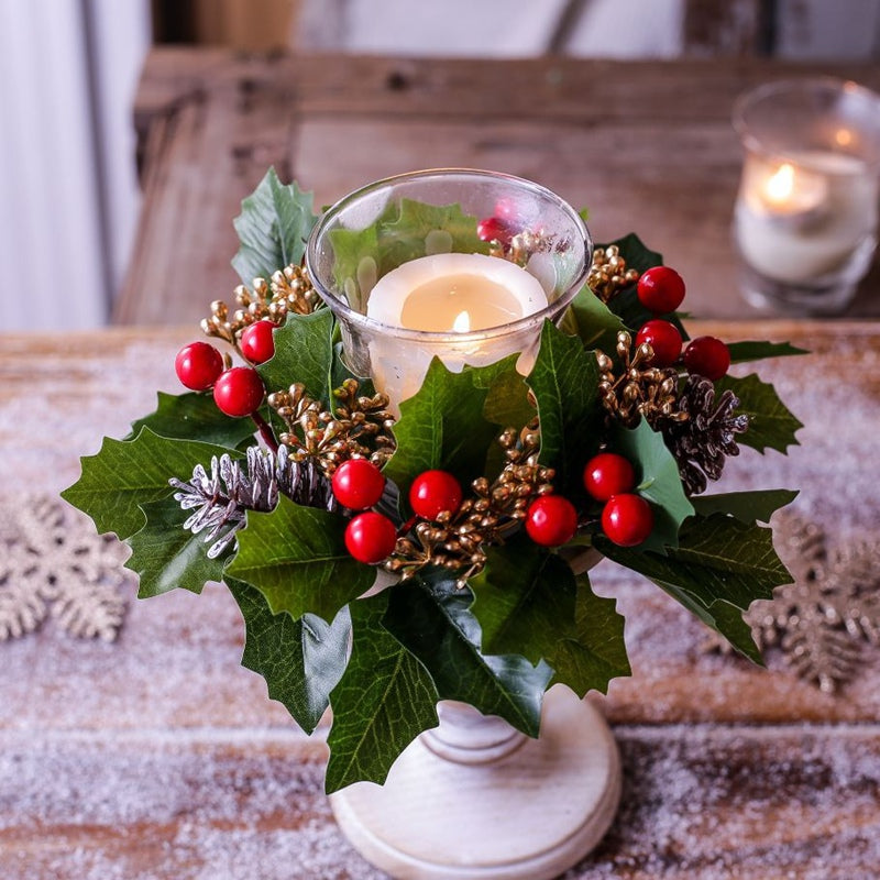 Artificial holly candle ring placed around glass candle holder, red berries and pinecones for rustic Christmas table decor