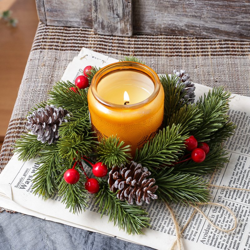 Festive candle ring with pine needles, red berries and frosted pinecones around lit amber candle, placed on vintage newspaper paper