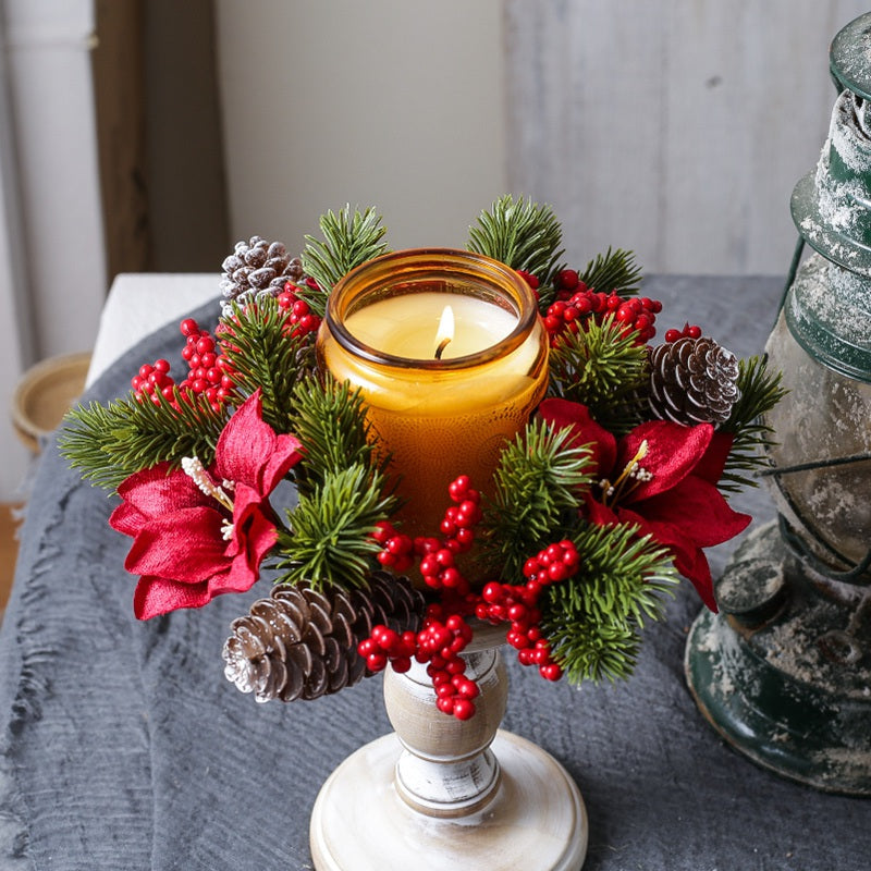 Christmas candle ring with poinsettia flowers, red berries and pine cones surrounding amber candle jar on distressed white wooden candlestick