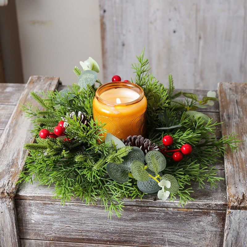 Christmas candle ring with artificial cypress, eucalyptus leaves, pine cones and red berries with amber candle on rustic wooden table