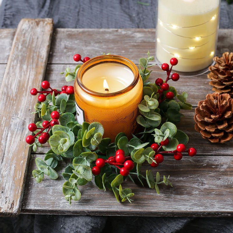 Eucalyptus red berry candle ring with lit amber candle on rustic wooden board with pine cones