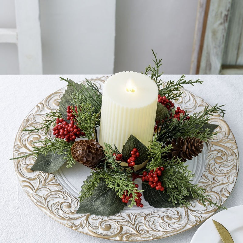 Christmas candle ring with pine cones, red berries and cypress leaves around ribbed pillar candle on textured white table with ornate plate