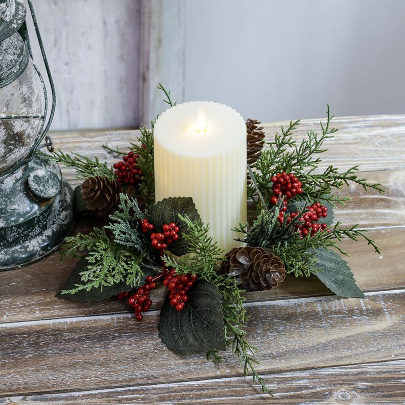 Christmas candle ring with cypress greenery, red berries and pinecones around ribbed pillar candle on rustic wooden table with vintage lantern