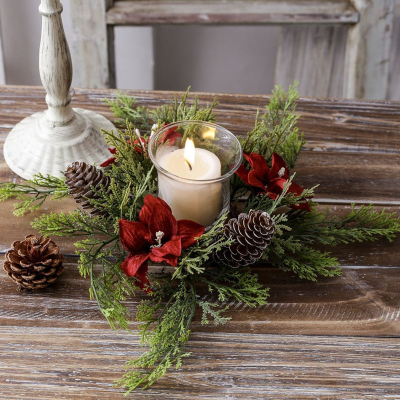 Red poinsettia Christmas candle ring with frosted pinecones and cypress foliage surrounding glass candle holder on weathered wooden table