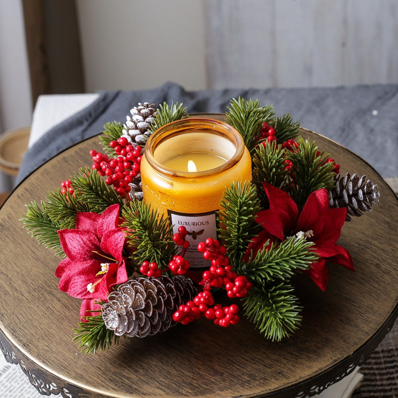 Small christmas candle wreath holder with red poinsettia, berries and pine cones around amber jar candle on rustic round wooden table