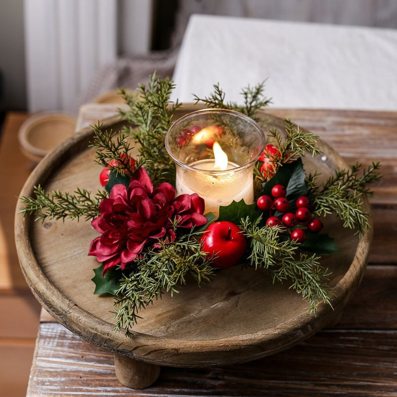 Christmas candle ring with burgundy dahlia, red berries, apples and evergreen sprigs, lit candle placed on round rustic wooden tray