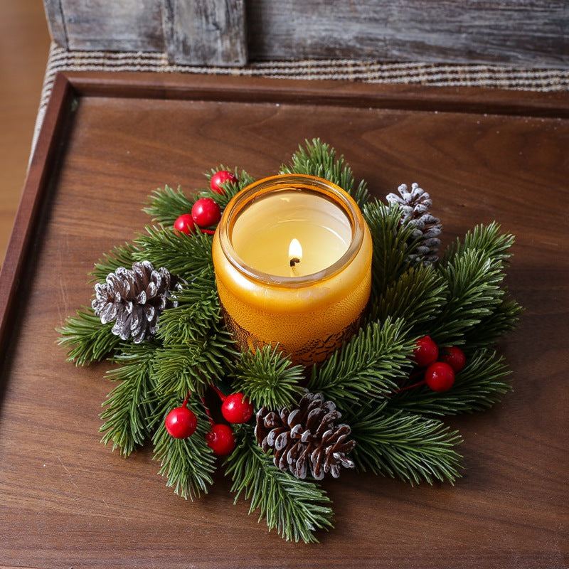Christmas candle ring with pine sprigs, red berries and frosted pinecones surrounding lit amber candle on dark wooden tray
