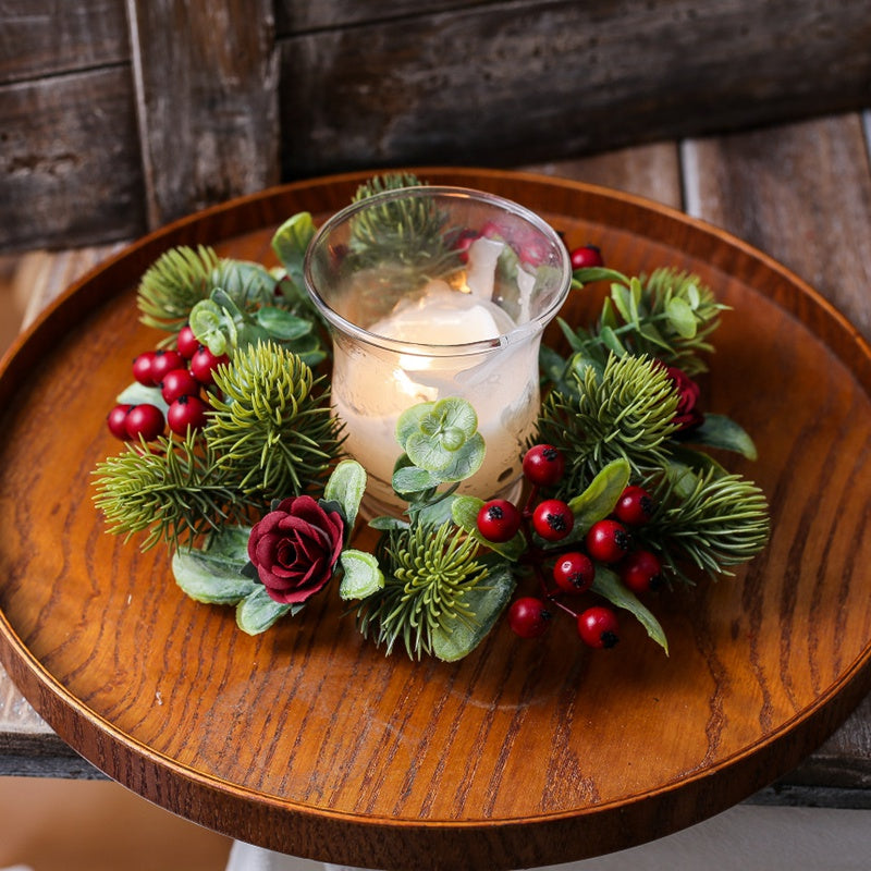 Artificial christmas candle wreath with red berries, burgundy roses and eucalyptus around glass candle on round wooden tray