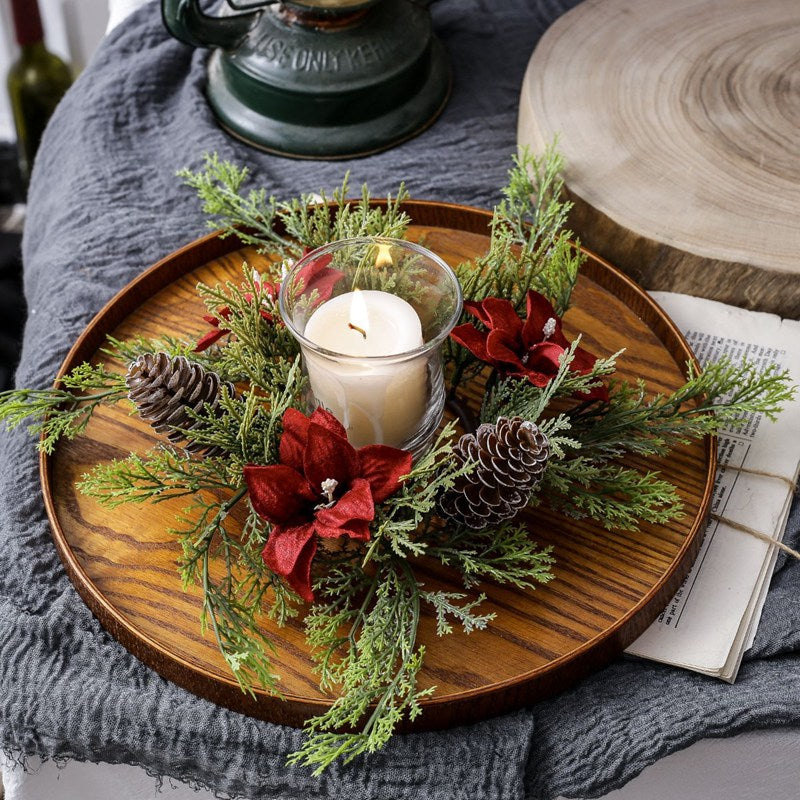 Artificial Christmas candle ring featuring red poinsettia, pine cones and cypress sprigs with glass candle cup on rustic wooden tray