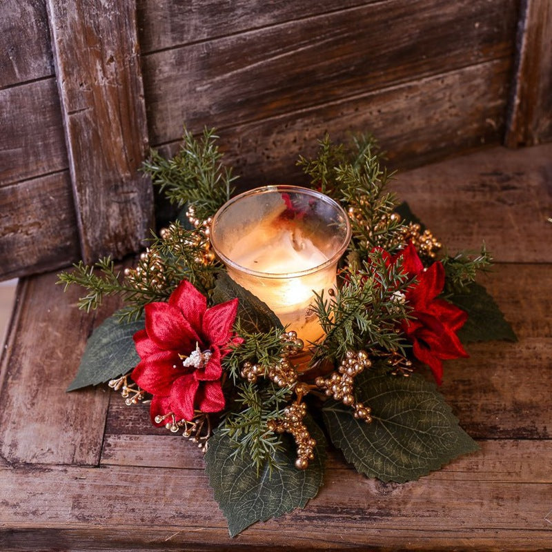 Artificial poinsettia candle ring with gold berries and cedar sprigs encircling lit glass candle on rustic wooden table