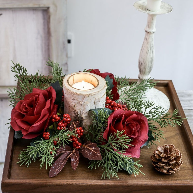 Holiday candle ring featuring burgundy roses, red berries and cypress foliage around birch candle on dark wooden tray, rustic Christmas table decor