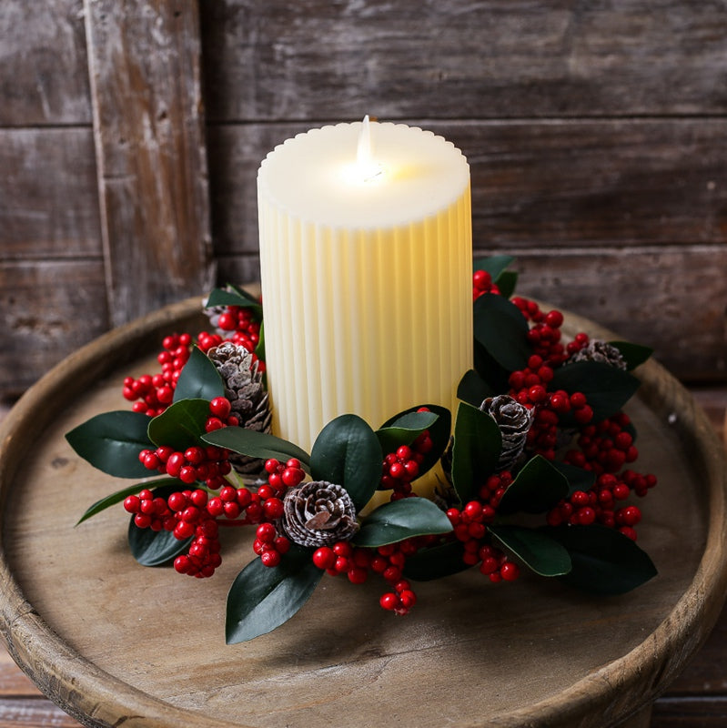 Christmas candle wreath holder with red berries and pine cones encircling fluted pillar candle on round rustic wooden tray